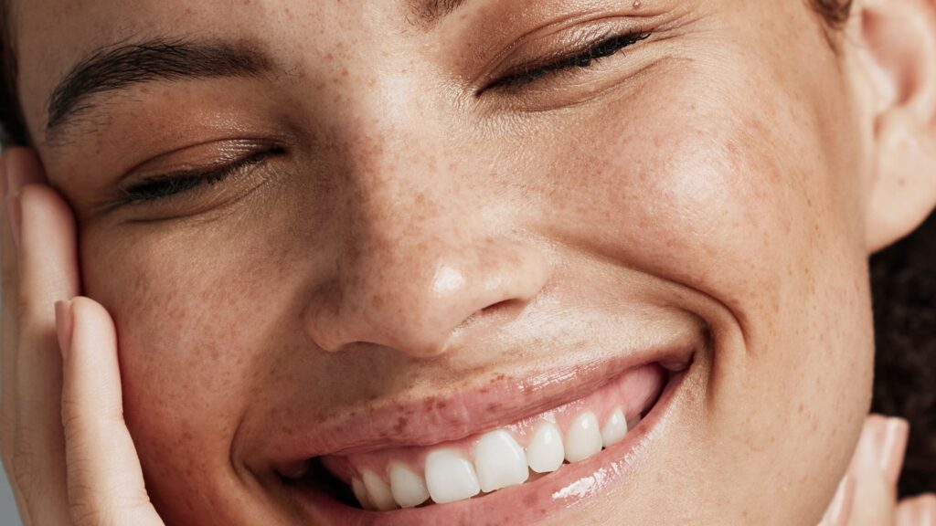 Close-up of a person with closed eyes and a big, joyful smile, gently touching their cheek. The Esse Sensitive Skin Trial / Travel Set brings out the natural glow in their freckles and enhances their skin texture.