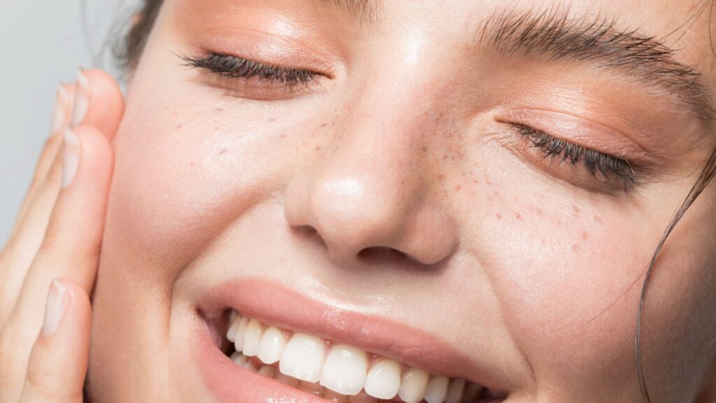 Close-up of a person with closed eyes and radiant, clear skin, thanks to the Esse Oily / Combination / Normal Trial / Travel Set. They are smiling broadly, revealing white teeth, and gently touching their cheek with one hand. They have light makeup with a natural look, including soft eyeshadow and light blush.