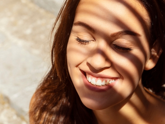 A woman with long brown hair smiles with her eyes closed as sunlight filters through blinds, casting striped shadows on her face.